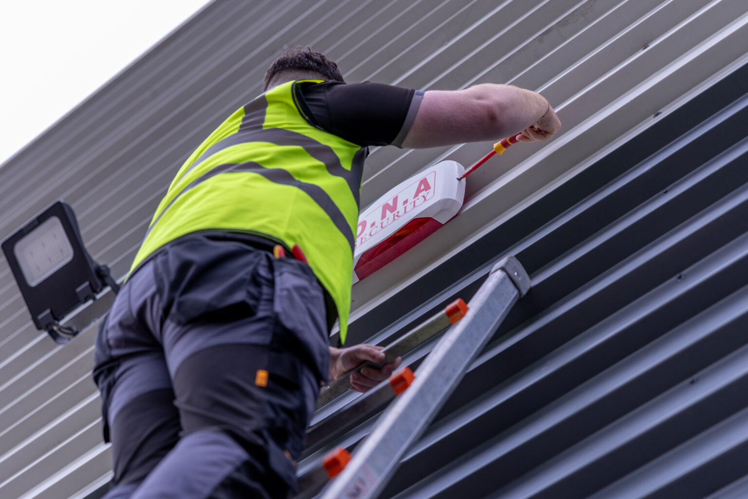 A DNA Security engineer on a ladder fitting an external alarm sounder to the wall of a commercial unit
